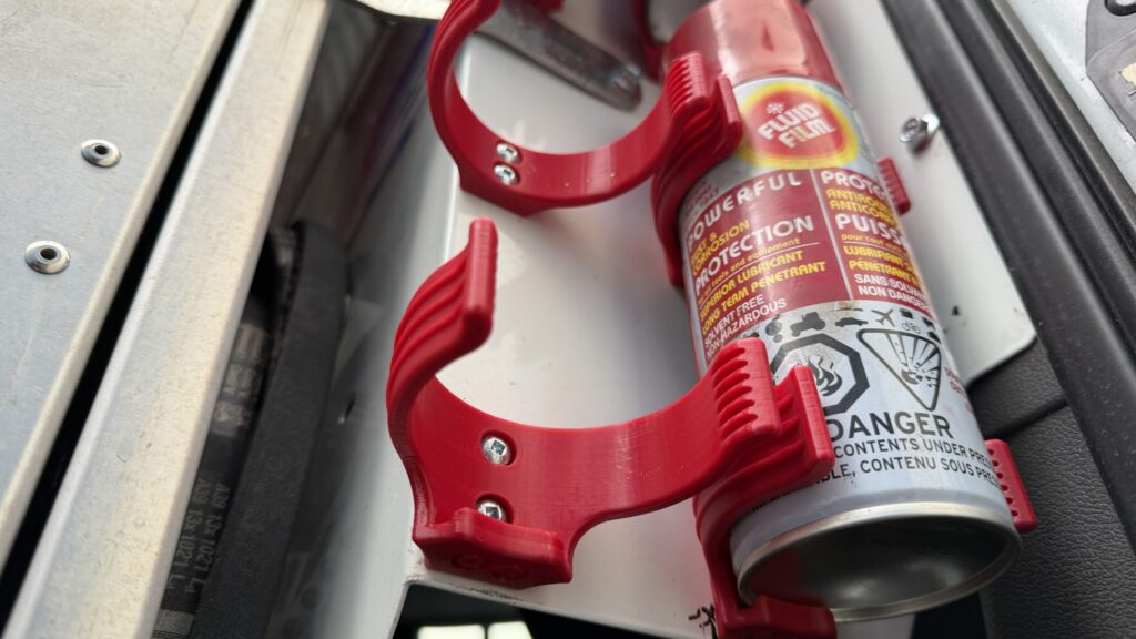 A close-up of a red 3D printed aerosol can holder mounted on a white metal surface inside a work van, holding a spray can.
