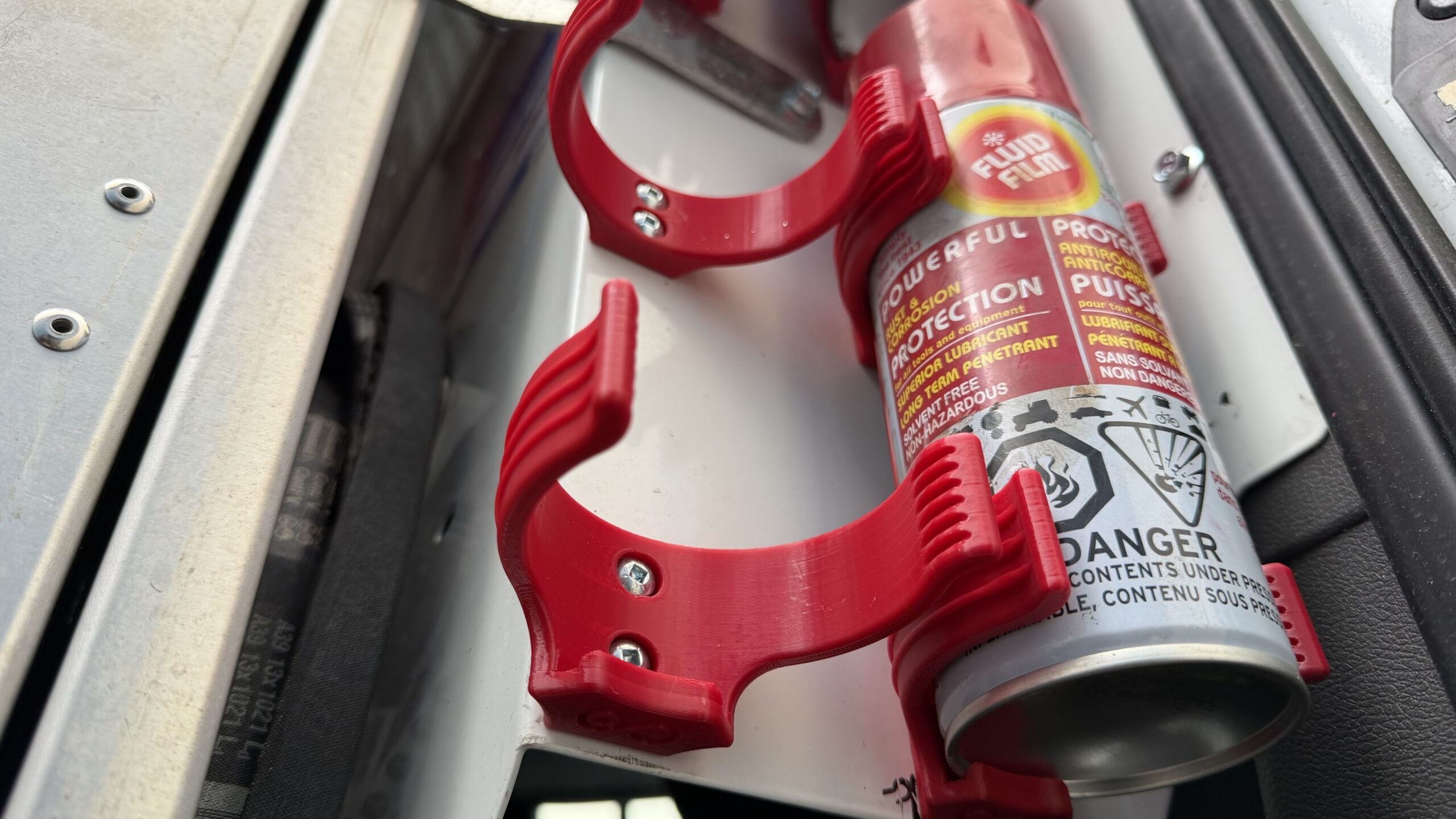 A close-up of a red 3D printed aerosol can holder mounted on a white metal surface inside a work van, holding a spray can.