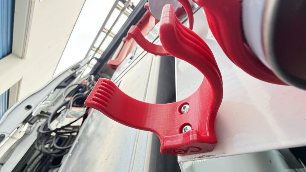 Low angle close-up of a red 3D printed spray can bracket mounted with two screws inside a work vehicle.