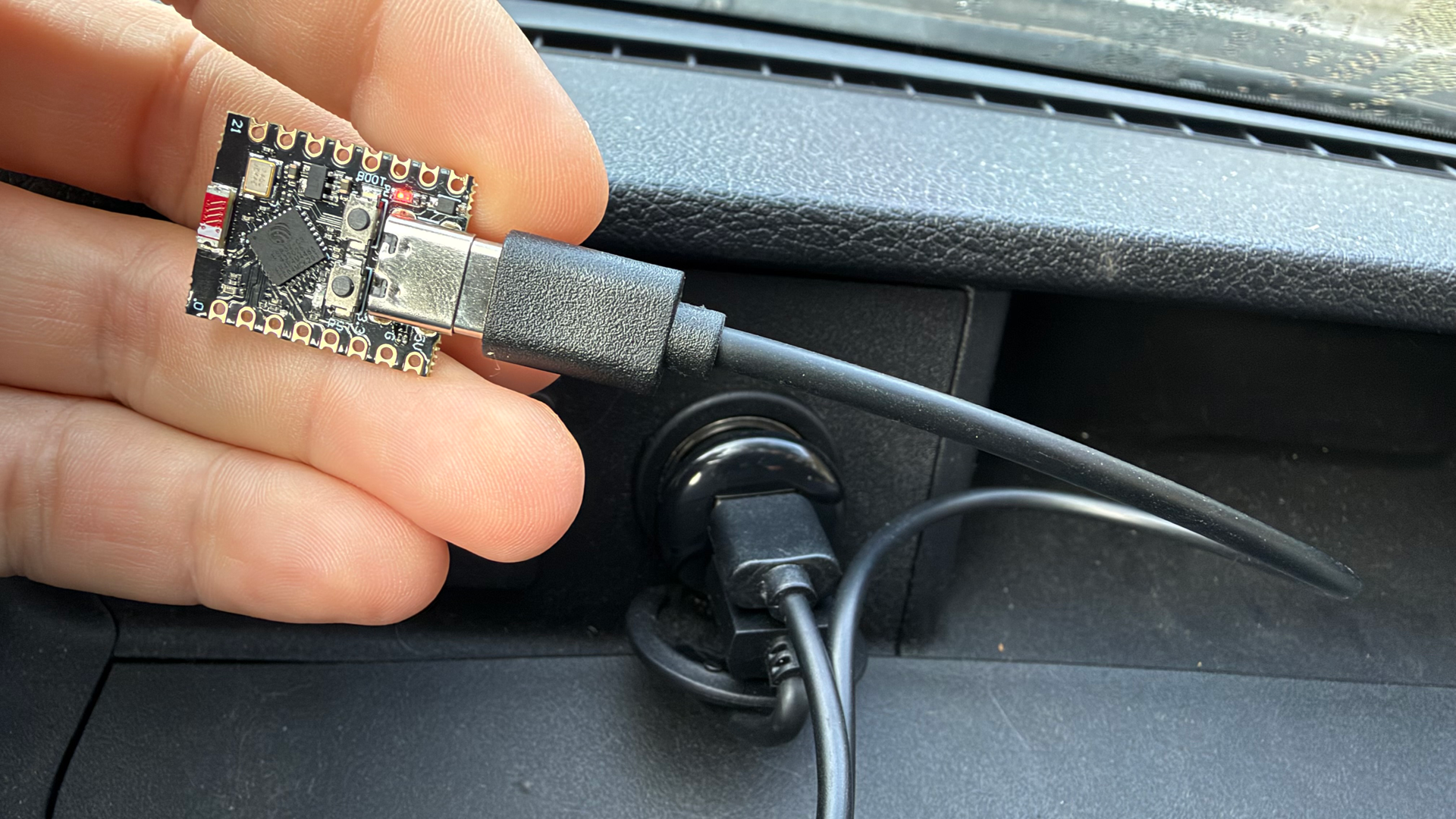 A hand holds a small ESP32 microcontroller board plugged into a USB cable inside a car.