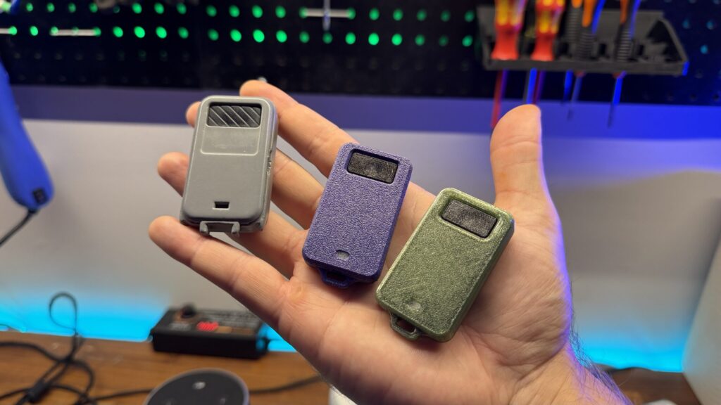 A hand holding three different colored 3D printed garage door remote keyfob casings against a workbench background.