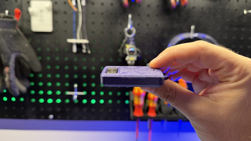 A person holds a small, purple 3D printed garage door remote keyfob housing in their hand in front of a pegboard.