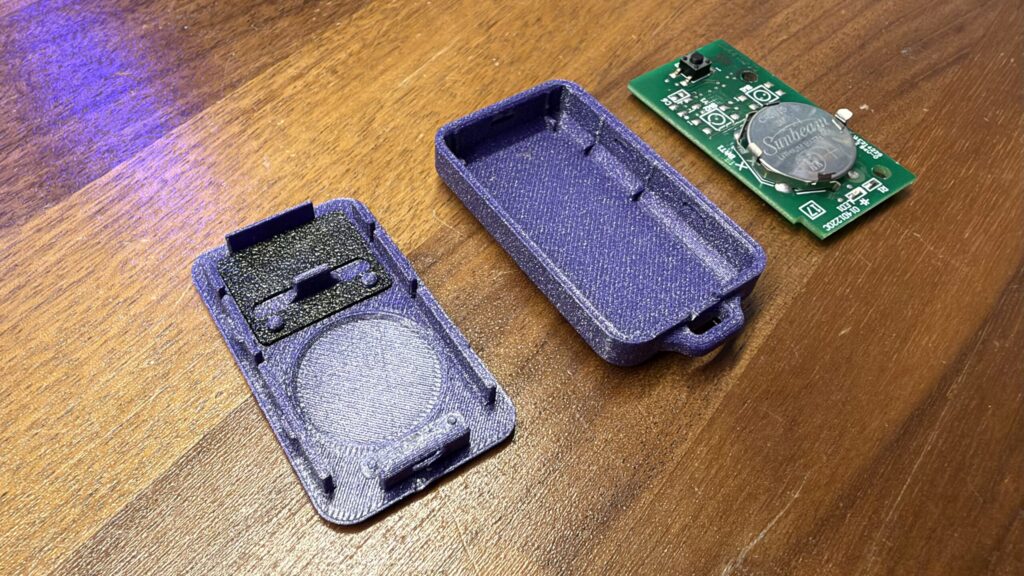 Disassembled purple 3D printed keyfob housing components and a green circuit board on a wooden table.