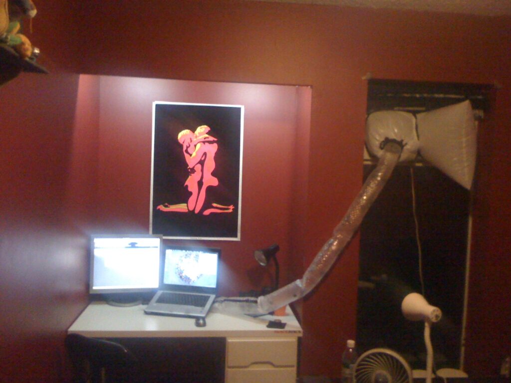 A laptop and monitor sit on a desk in a red room, connected to a DIY air conditioning duct made of garbage bags and plastic bubble wrap